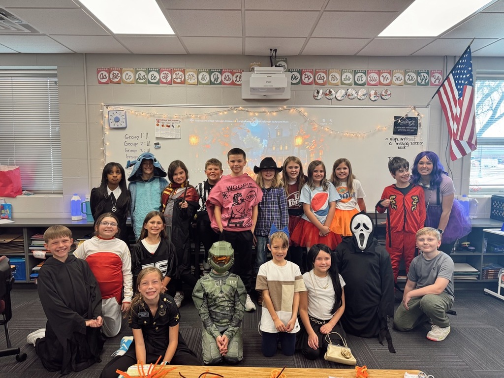 Group of children and their teacher in front of their classroom posing for a picture with most wearing Halloween costumes.