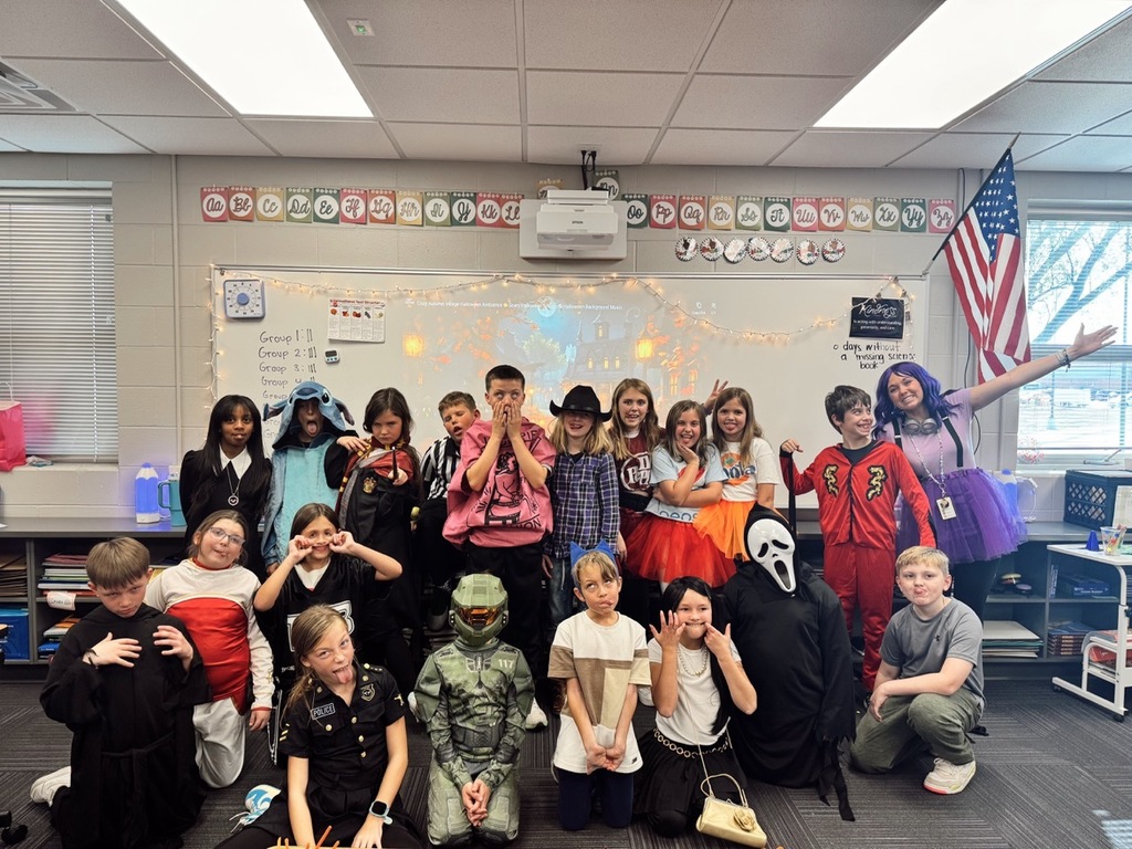 Group of children and their teacher in front of their classroom posing and making silly faces for a picture with most wearing Halloween costumes.
