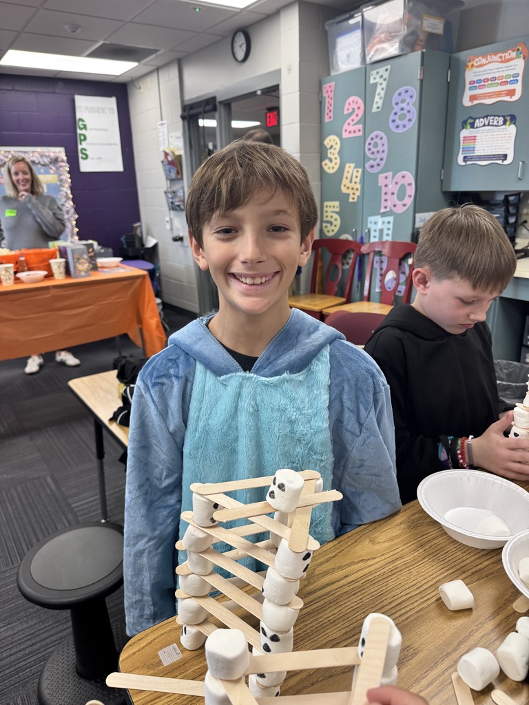 Student smiling while standing in front of a table with a craft of popsicle sticks and marshmallows.