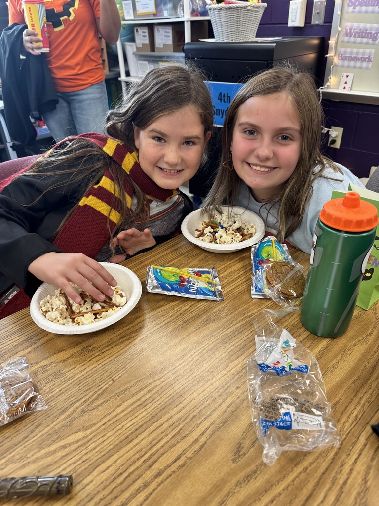 Two girls sitting at a table with a bowl full of popcorn, pretzels, and juice boxes on the table.