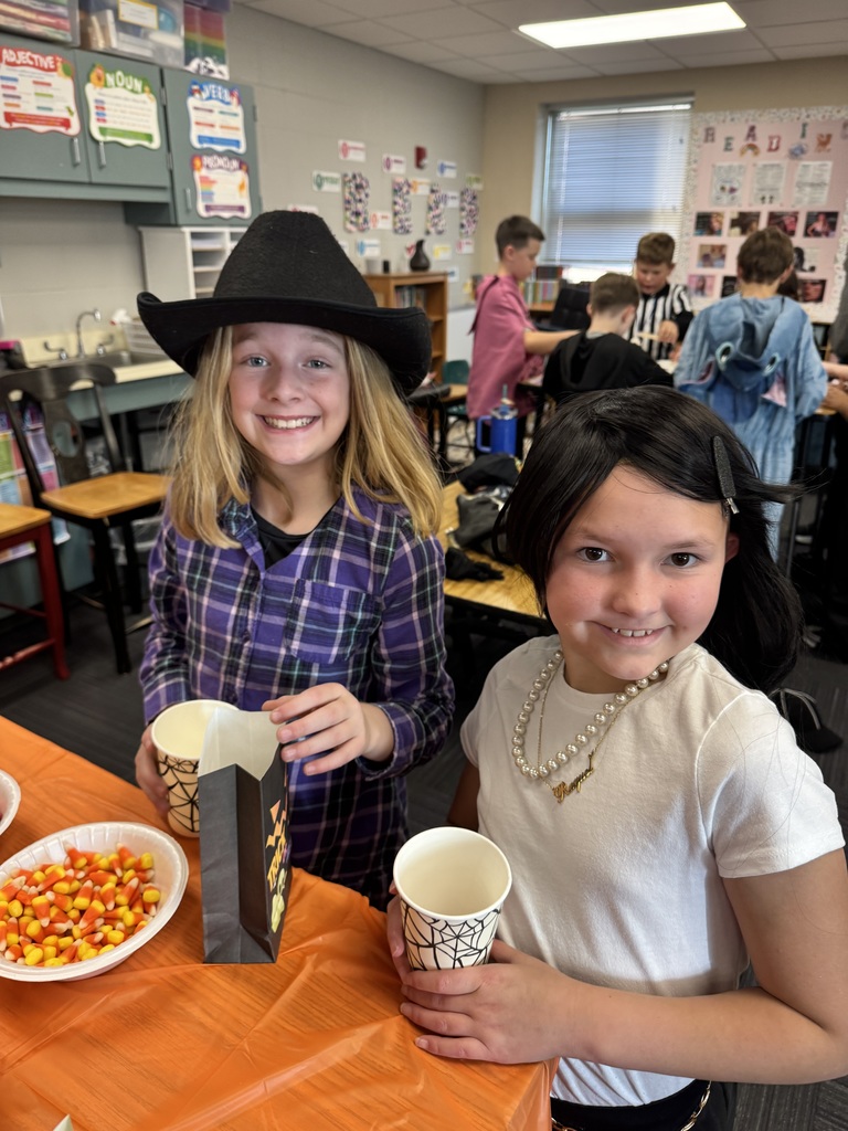 Two girls, one wearing a cowboy hat and one wearing a black wig filling up a paper bag with popcorn and candy corn.