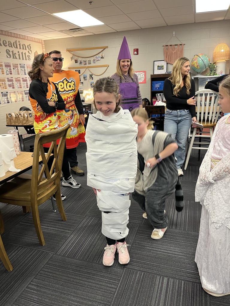 Girl standing in a classroom while a student wraps her in toilet paper to look like a skeleton.