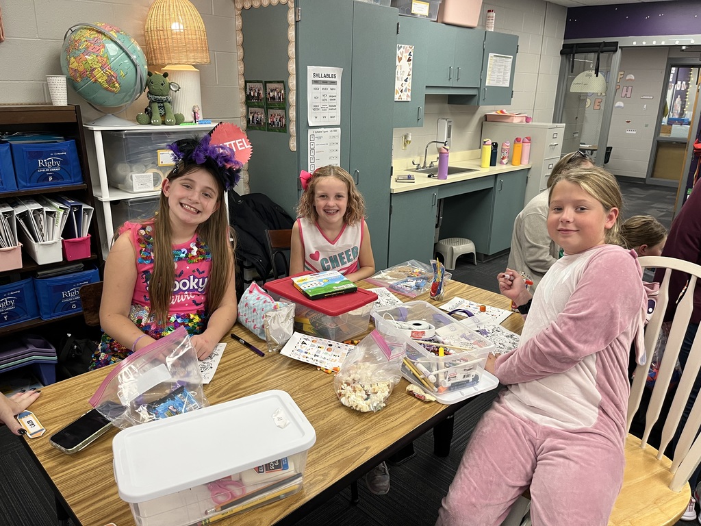 Four girls sitting at a table in a classroom with art supplies visible and a bag of popcorn and candy sitting on it.