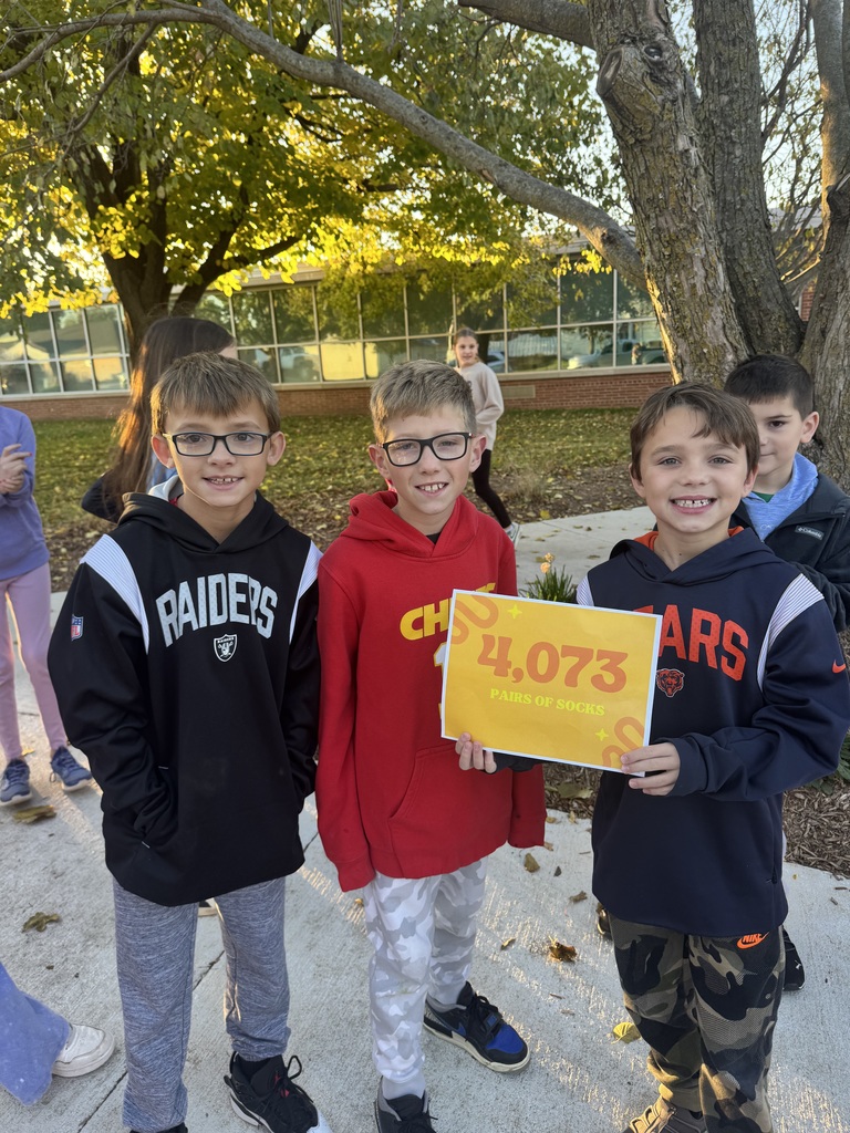 Three boys standing outside on a sidewalk while wearing hoodies, one boy is holding a sign that shows the number 4,073