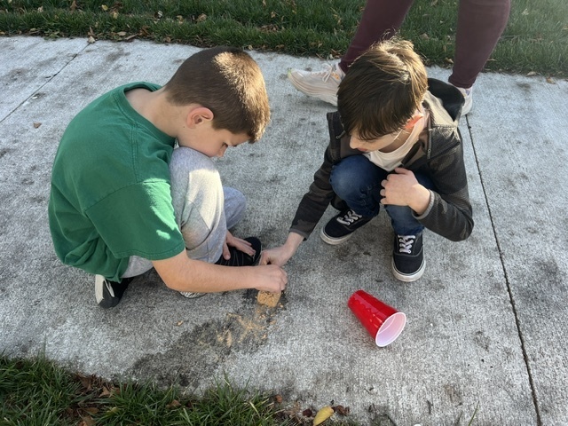 two boys sitting on a sidewalk digging through a rock with a red cup visible and water on the sidewalk.