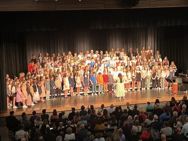 Photo of middle school choir students performing on stage with their music teacher in front directing them.