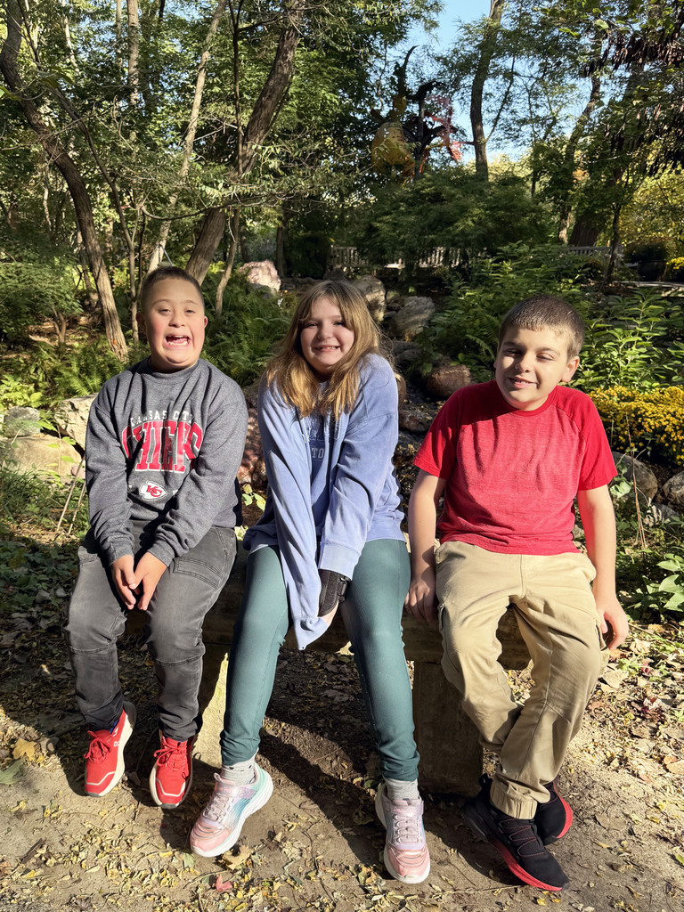 three students sitting on the side of a dirt path with gardens and water behind them