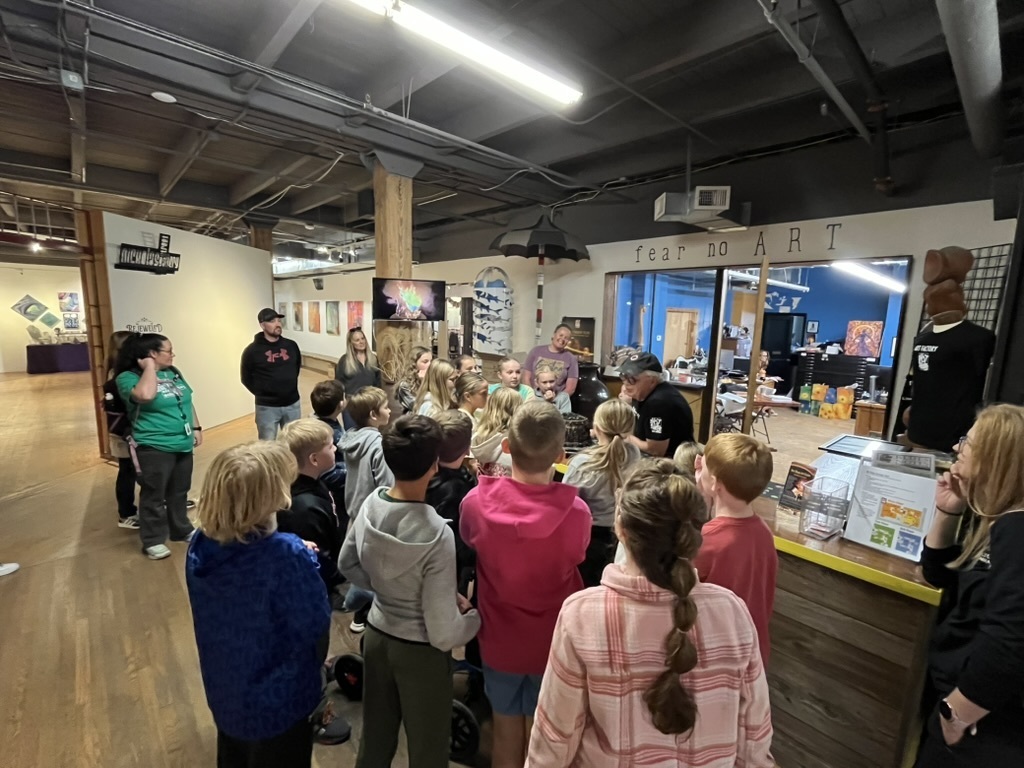 Group of students and adults gathered around a man in an art studio who appears to be showing them a pot.