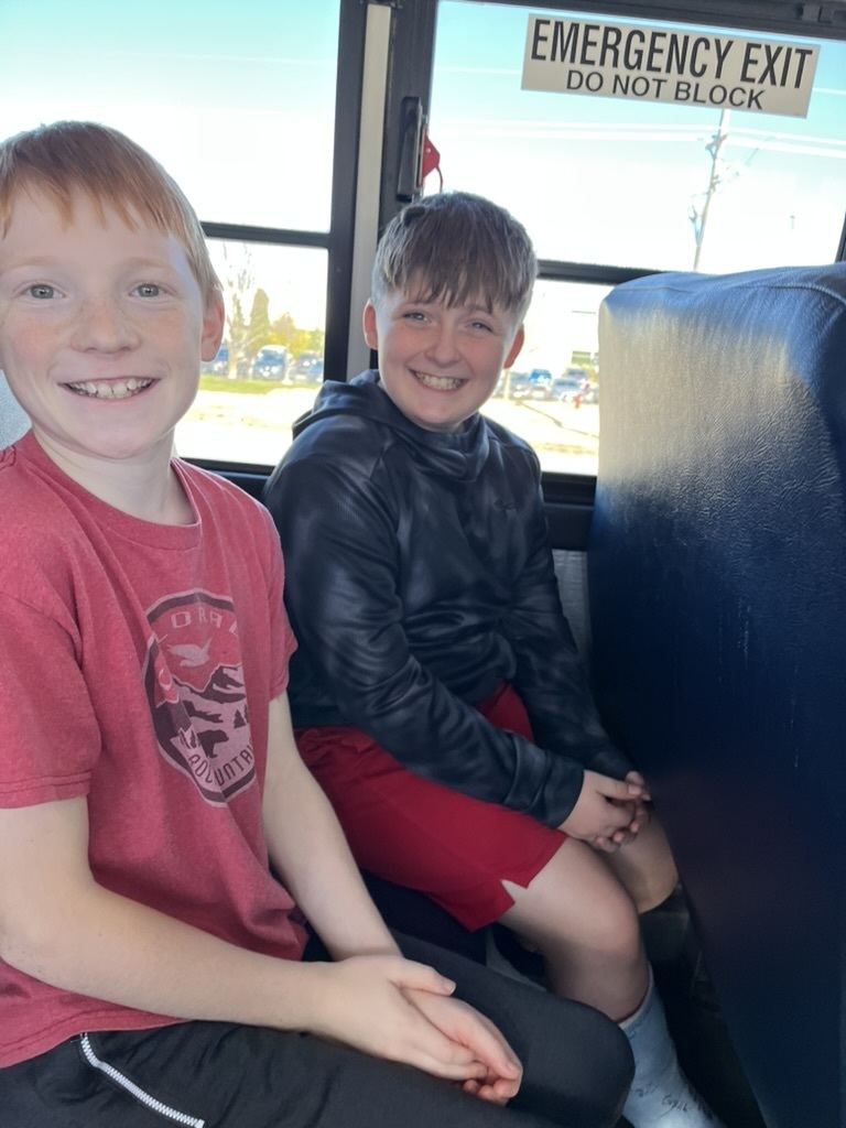 Two boys sitting on a bus seat smiling at the camera.