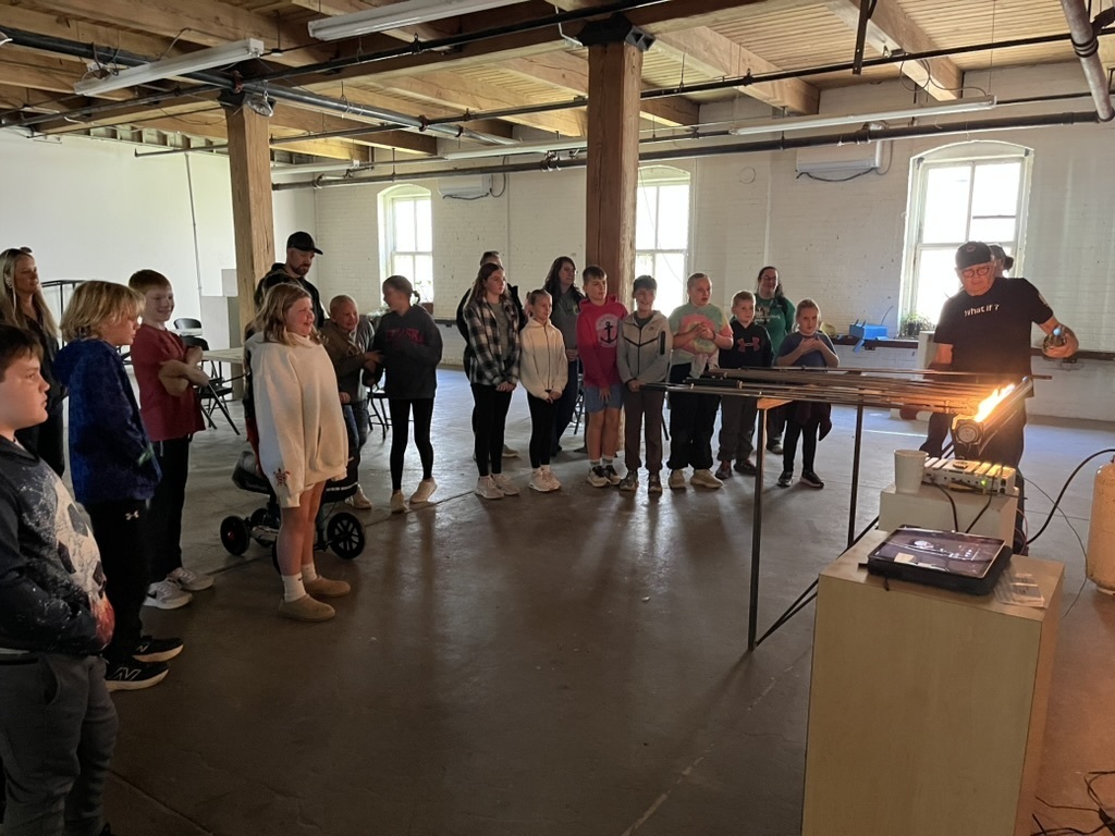 Group of students and adults standing around a man who is showing them an art instrument that involves flames.