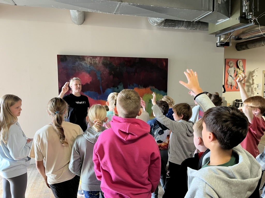 Group of students standing around an adult in an art studio with some students having their hands raised.