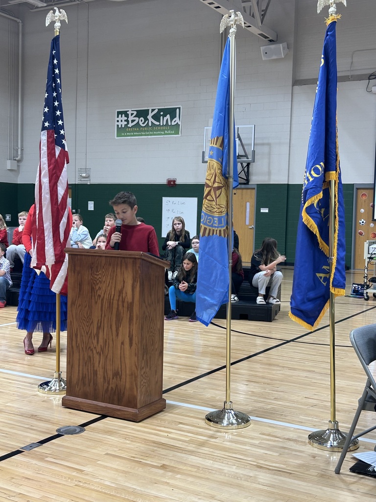 Student stands at a podium reading a speech with his teacher standing next to him.  Flags are present with students sitting on risers behind them.