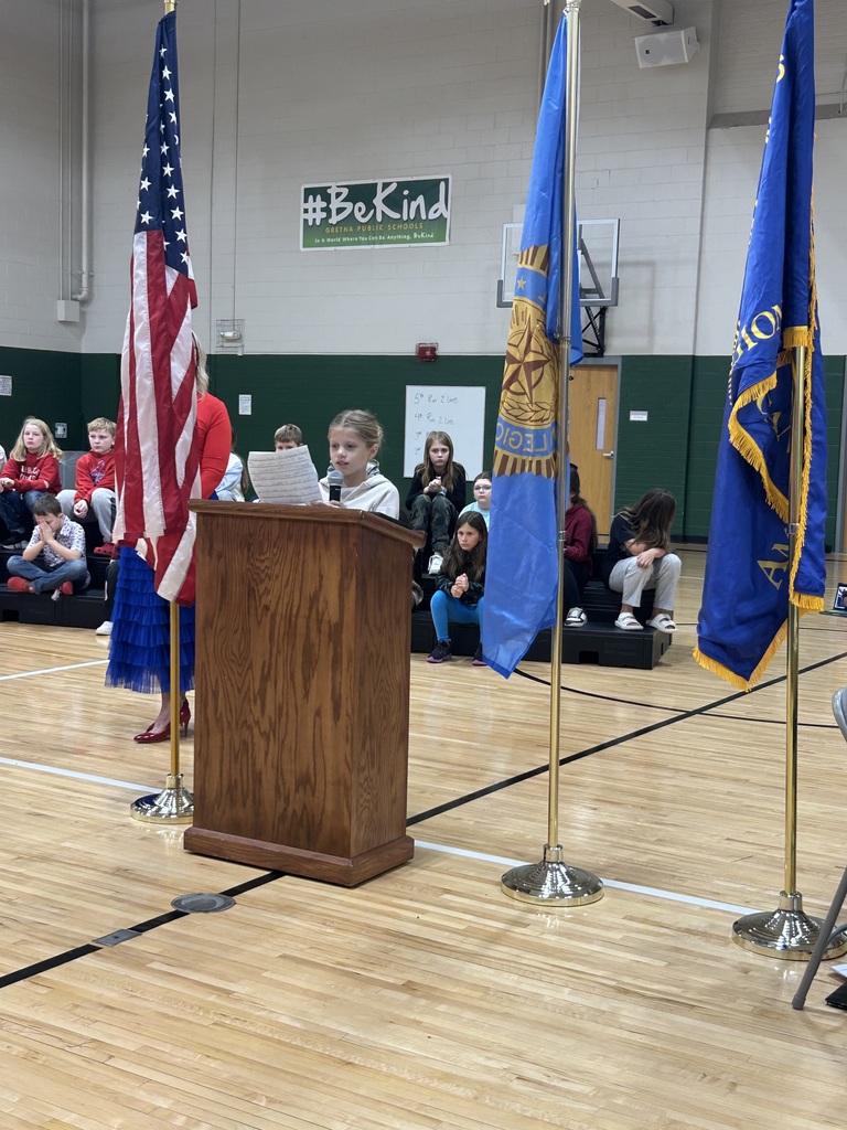 Student stands at a podium reading a speech with her teacher standing next to her.  Flags are present with students sitting on risers behind them.