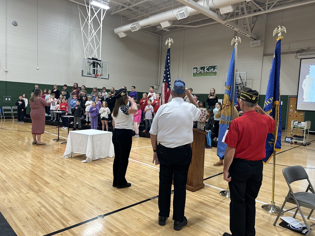 Group of children with hands on their hearts singing the National Anthem with members of the military saluting in a gym.