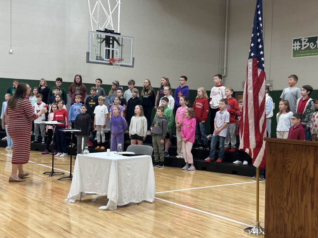 Group of students stand on risers singing while a teacher conducts in front of them with the American Flag visible.