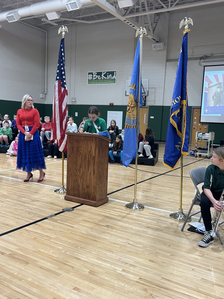 Student stands at a podium reading a speech with her teacher standing next to her.  Flags are present with students sitting on risers behind them.