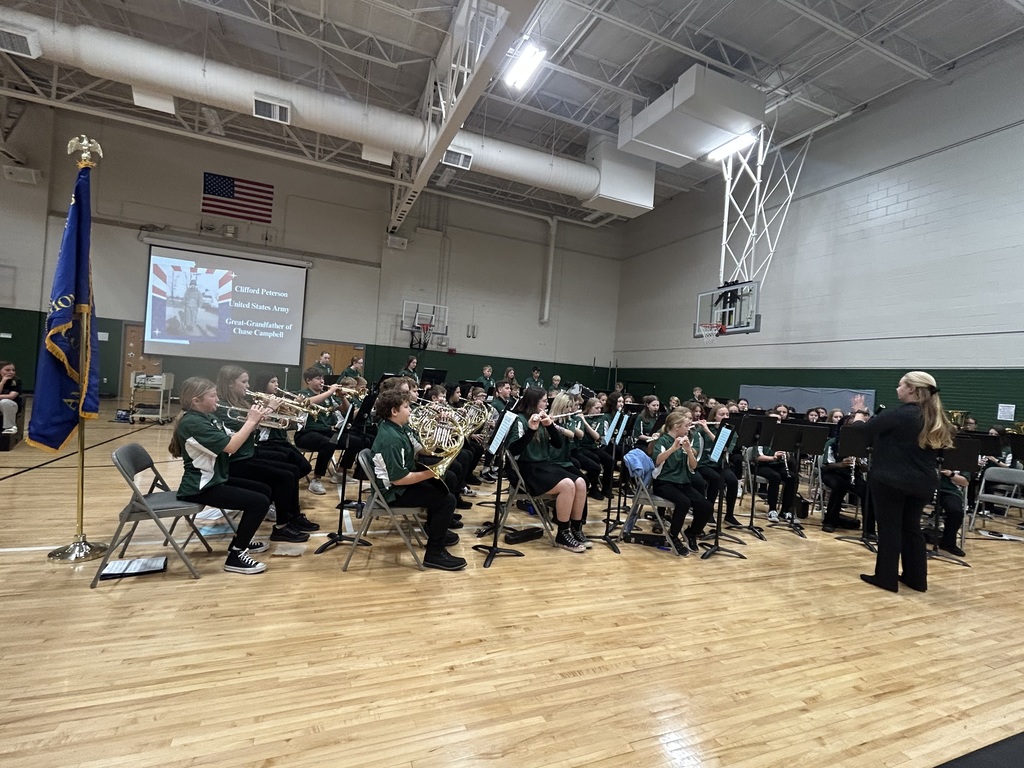 Members of a middle school band wearing green shirts playing their instruments with their teacher in front and a flag visible.