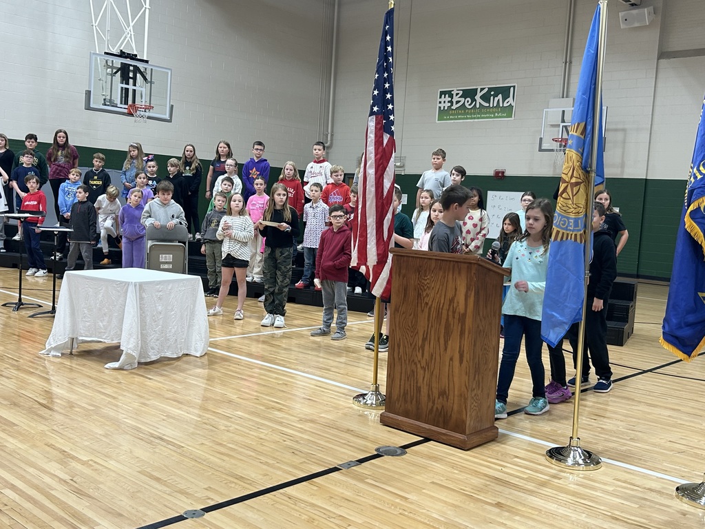 Group of elementary students standing on risers while some stand in front of a podium with flags visible.