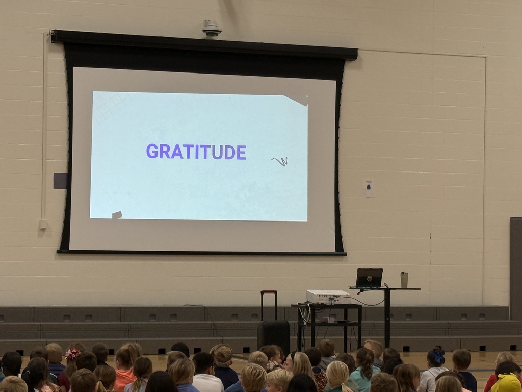Students sitting on the gym floor looking at the screen with the word "Gratitude"