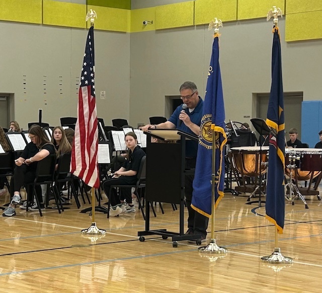 Photo of Major Jim Oliver speaking at the Veteran's Day assembly with flags on each side of him.