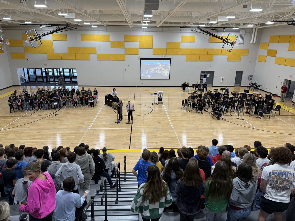 Photo of Mr. Gross speaking in the gym at the assembly with the band, choir, and other students watching.