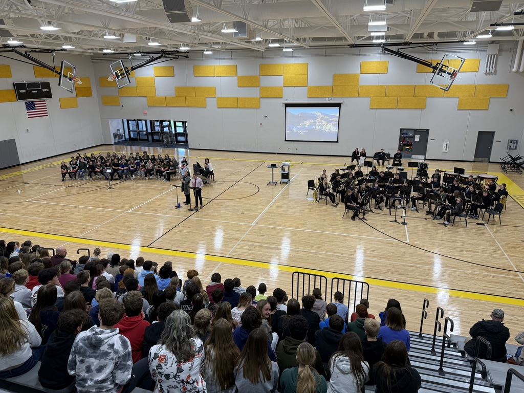 Photo of Mr. Gross speaking in the gym at the assembly with the band, choir, and other students watching.