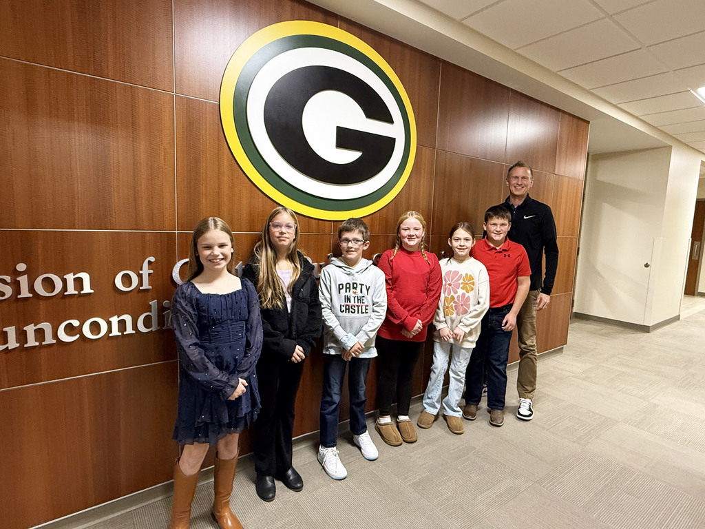 six students and teacher standing in front of the Gretna public school "G" logo and mission statement wall with teacher