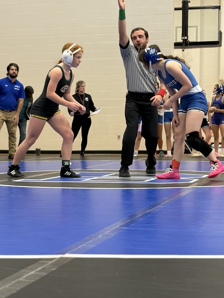 Two female wrestlers face each other. A male ref stands between them with his hand up in the air. 
