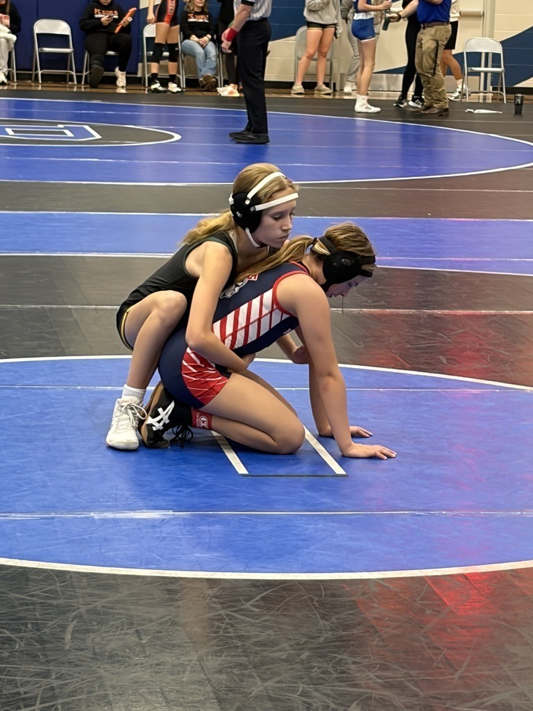 Two female wrestlers are on the mat in the start position (one is on her hands and knees and the other is behind her). 