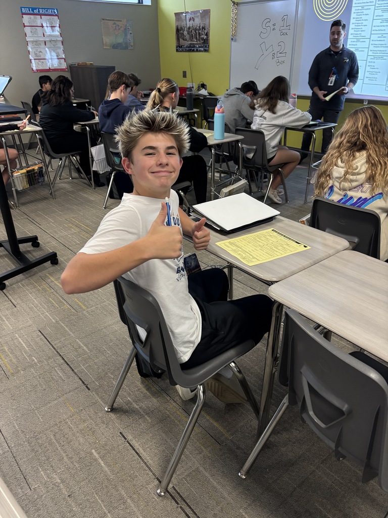 Photo of a student giving the camera a thumbs up during the counselor's presentation. The counselor is standing at the front of the room looking out at the students.