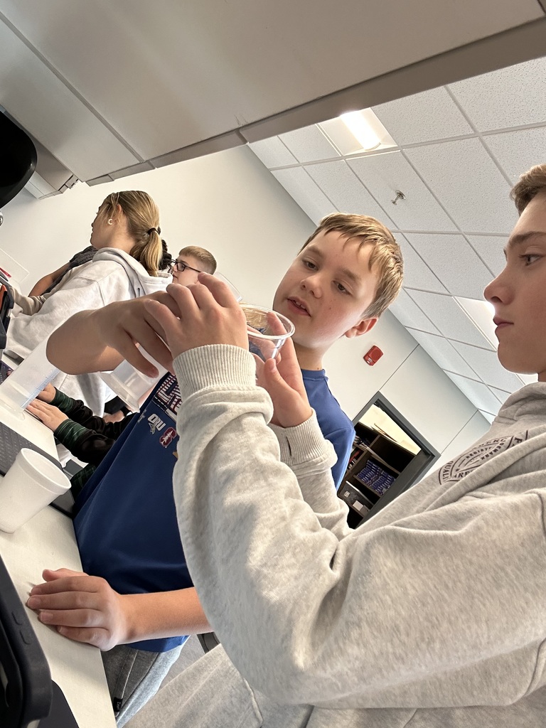 Two students are holding cups and beakers to measure. There are other students working in the background. 