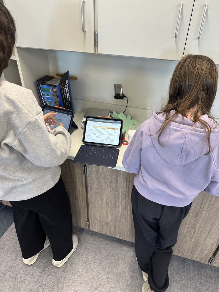 Two female students are standing at a counter with their backs to the camera. They have an iPad between them. 