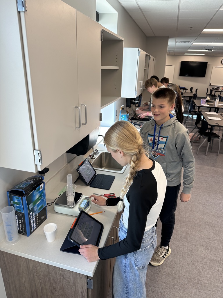 Students are standing at a long counter, working on measuring weights and recording data on their iPads. 
