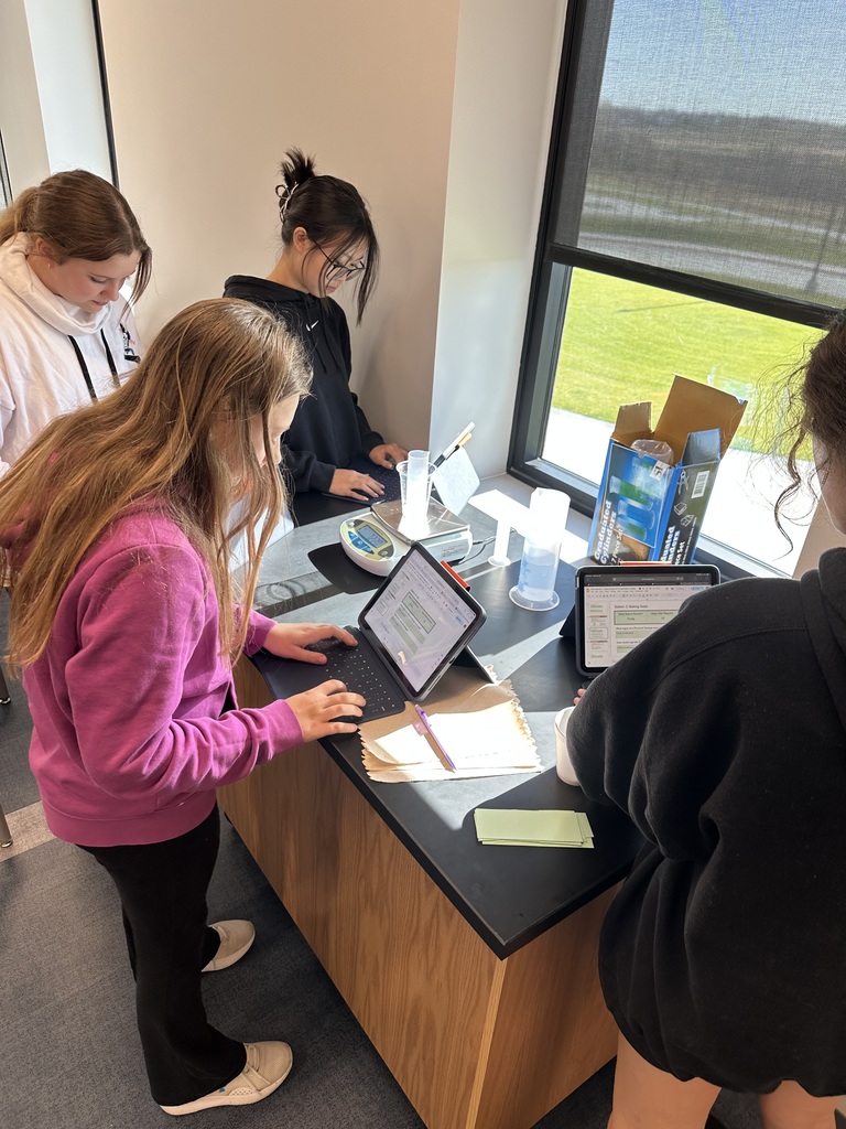 A group of 4 female students are working at a black desk. One is typing on an iPad while the others are looking at their materials. 