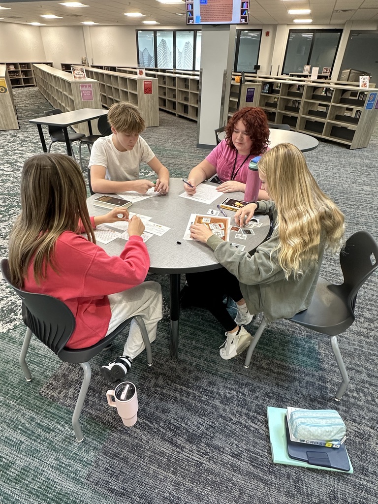 Four students sit at a table with various pieces of paper. They are all looking at the pages. 