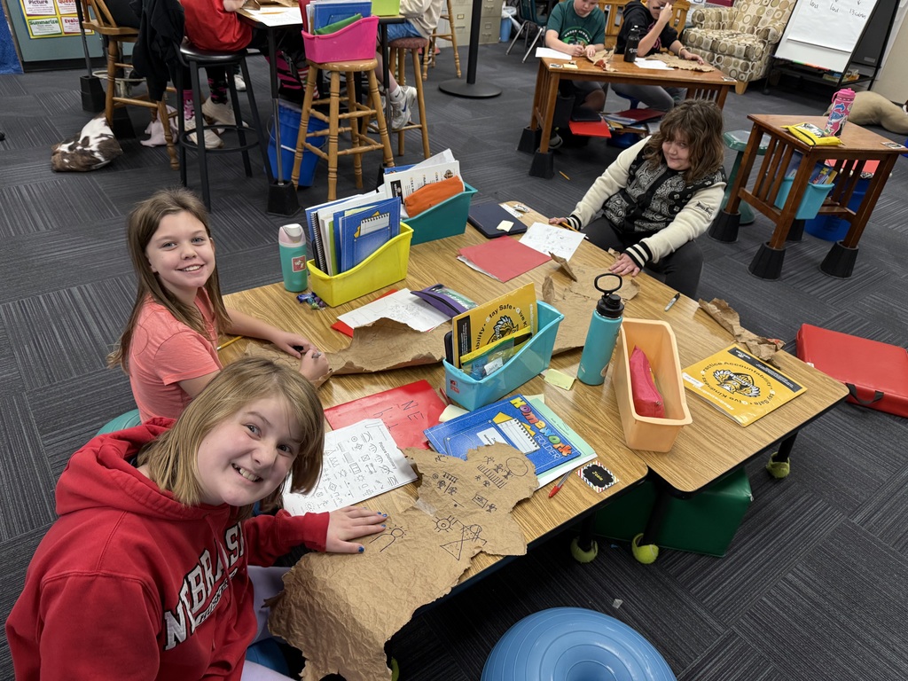 Three girls sitting at a table working on designs made out of paper bags with drawings on them that resemble buffalo hides.