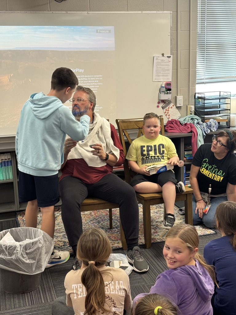 Man sitting on a chair getting his beard dyed blue with an adult and students watching.