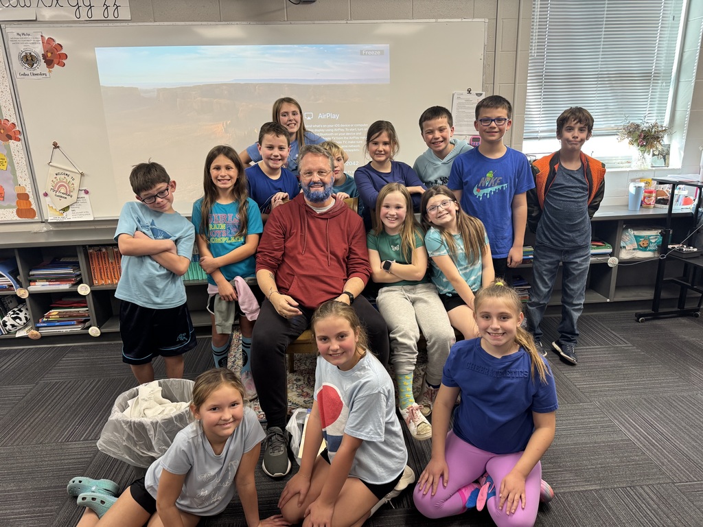 Group of students standing and sitting around a man in a chair with a blue beard.