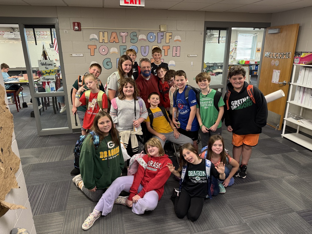 Group of students standing and sitting in a hallway around a man with a blue beard.