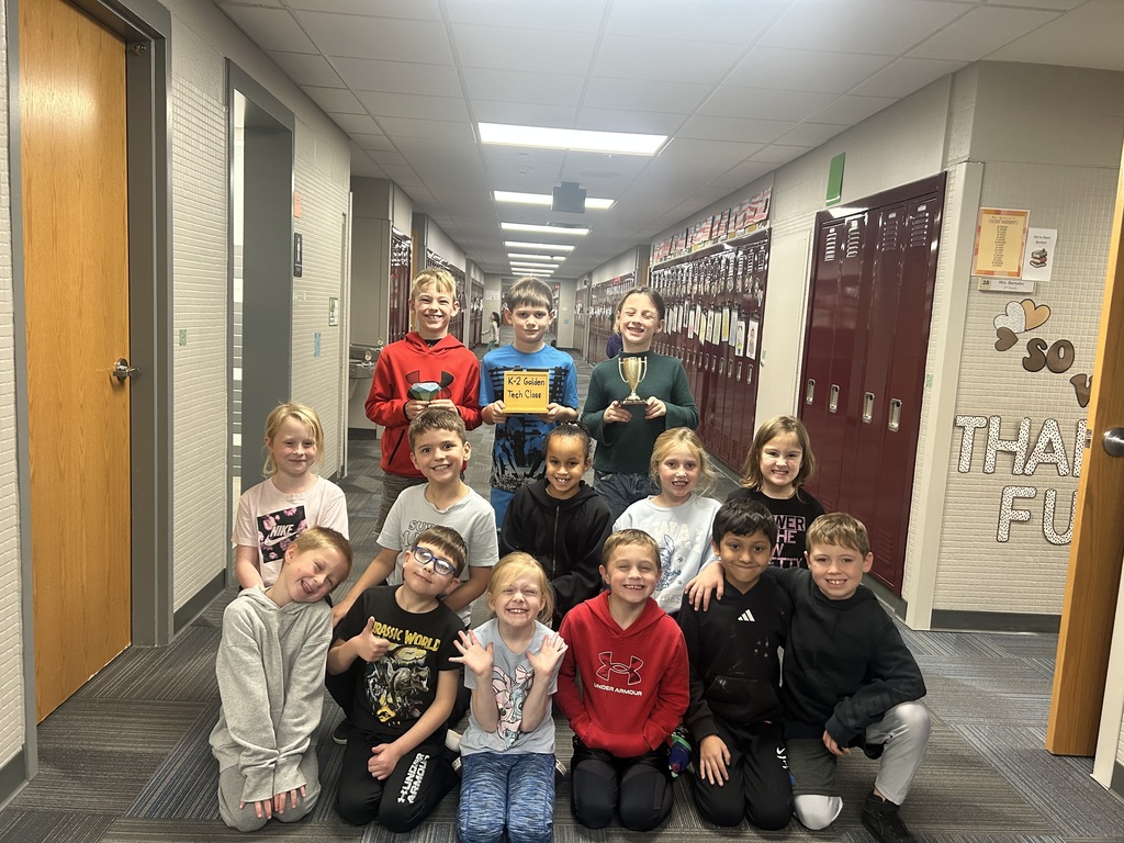 Group of second grade students, some standing, some kneeling, and some standing, holding awards in a school hallway.