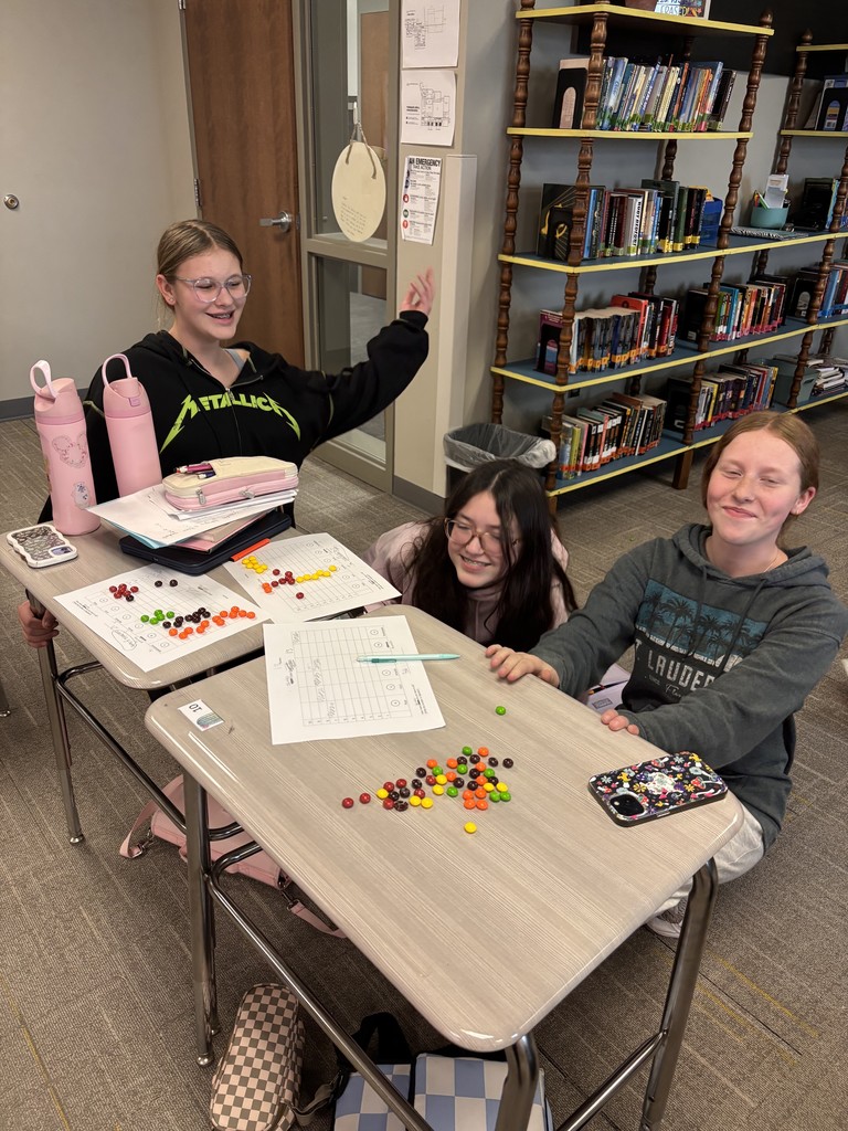 Photo of three students sitting at desks looking at the camera and smiling with Skittles and paper in front of them for their activity.