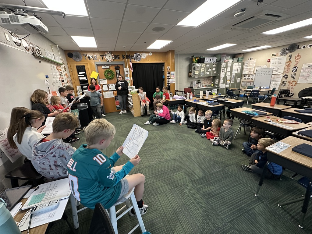 Group of students standing at the front of a classroom holding papers to read while other students sit on the floor around them.