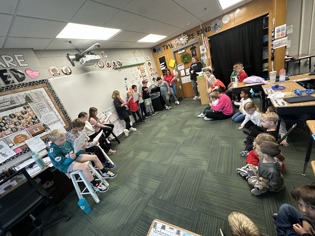 Group of students standing at the front of a classroom holding papers to read while other students sit on the floor around them.