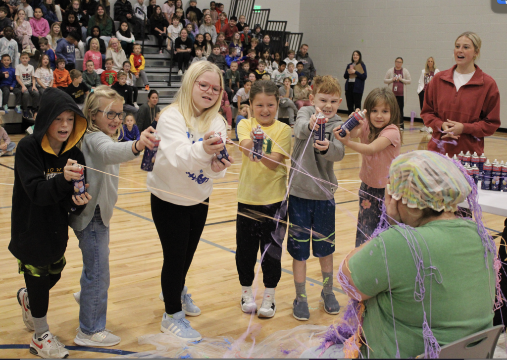 Mrs. Hardy covers her face as second graders spray silly string on her.