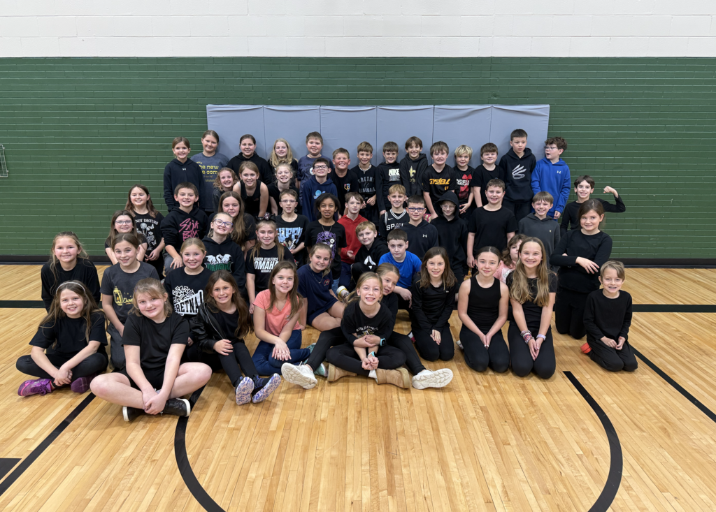 Group of students, many of them wearing black, sitting and standing for a photo in a gym with green walls.