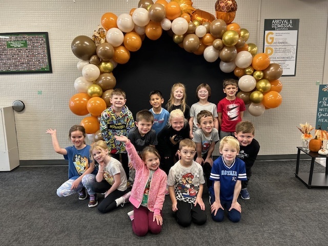 Group of students kneeling and standing around an arch made of Thanksgiving colored balloons.