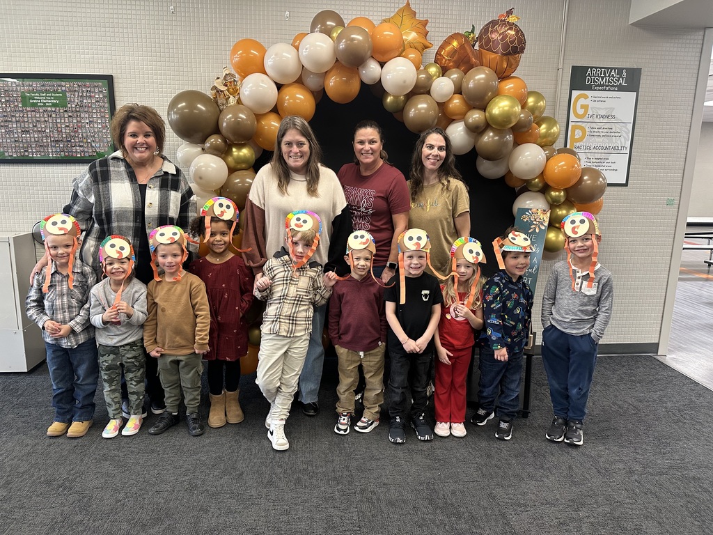 Group of preschool students wearing turkey hats with adults standing behind them in front of an arch made of Thanksgiving colored balloons.