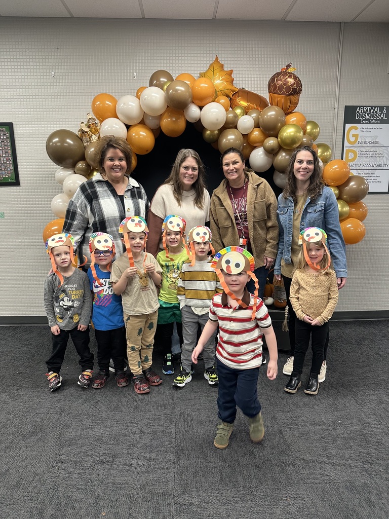 Group of preschool students wearing turkey hats with adults standing behind them in front of an arch made of Thanksgiving colored balloons.