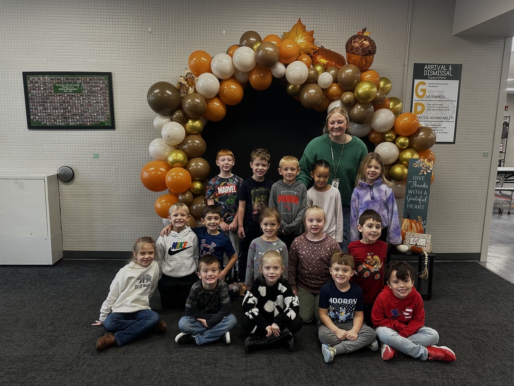 Group of students, some sitting, some kneeling, and some standing, along with their teacher in front of an arch made of Thanksgiving colored balloons.