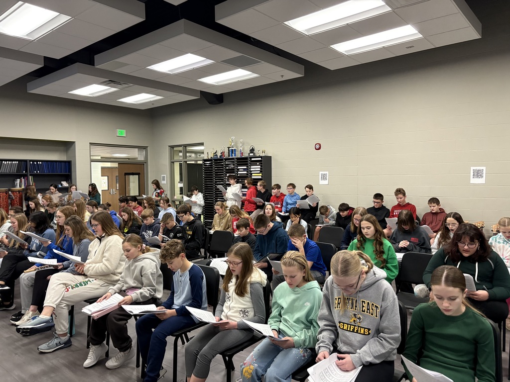 Photo of multiple rows of choir students sitting in chairs looking at sheet music to practice singining.